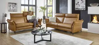 Zweiteilige Ledergarnitur in Cognacfarbe mit hoher Rückenlehne, Frau liest entspannt im hellen Wohnzimmer mit Kamin und moderner Einrichtung.